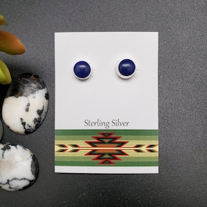 Peut inclure: Boucles d'oreilles clous en argent sterling avec des pierres rondes bleu foncé. Les boucles d'oreilles sont présentées sur une carte blanche avec l'inscription "Sterling Silver" et un motif de style sud-ouest. Fond gris foncé.