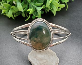 May include: A silver cuff bracelet featuring an oval, green and brown gemstone. The gemstone is set within a decorative, rope-style border. The bracelet has a simple, elegant design, suitable for everyday wear.