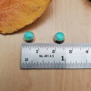 Puede incluir: Dos pendientes redondos de ópalo verde con engastes de plata, medidos con una regla. Los pendientes tienen un diámetro aproximado de 1,2 cm.