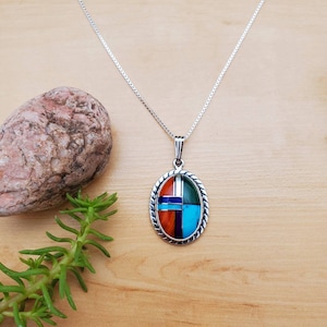 Pu&ograve; includere: Una collana con catena d'argento con un ciondolo ovale che presenta un design geometrico di pietre turchesi, rosse, verdi e blu incastonate.