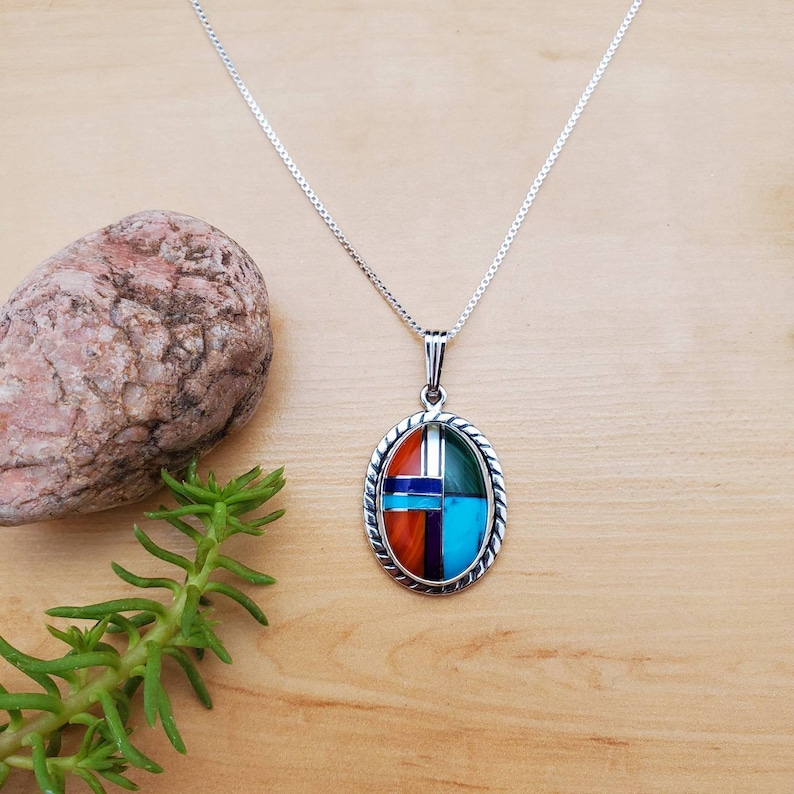 Pu&ograve; includere: Una collana con catena d'argento con un ciondolo ovale che presenta un design geometrico di pietre turchesi, rosse, verdi e blu incastonate.