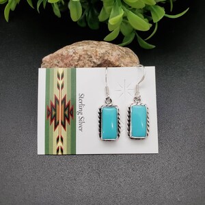 Puede incluir: Pendientes rectangulares turquesa con un borde plateado en forma de cuerda. Los pendientes se muestran en una tarjeta blanca con las palabras "Sterling Silver" y un diseño de inspiración nativa americana. El fondo es negro.