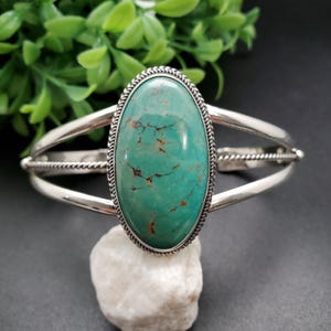 Könnte beinhalten: Ein silbernes Armreif-Armband mit einem großen, ovalen Türkisstein. Der Stein hat eine leuchtende türkisblaue Farbe mit braunen Adern. Das Armband hat ein schlichtes Design mit einem gedrehten Seildetail.
