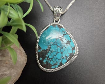 Può includere: Una collana con pendente caratterizzata da una grande pietra turchese irregolare con venature a matrice marrone scuro. La pietra è incastonata in una cornice d'argento con bordo a corda ritorta e una contromaglia decorativa.