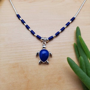 Può includere: Una collana d'argento con un ciondolo a forma di tartaruga in lapislazzuli blu. La collana è composta da perline d'argento e perline di vetro blu.