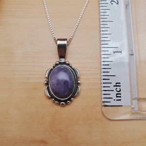 May include: A silver chain necklace with a purple oval gemstone pendant. The pendant is set in a silver frame with a decorative design. The necklace is measured against a ruler.