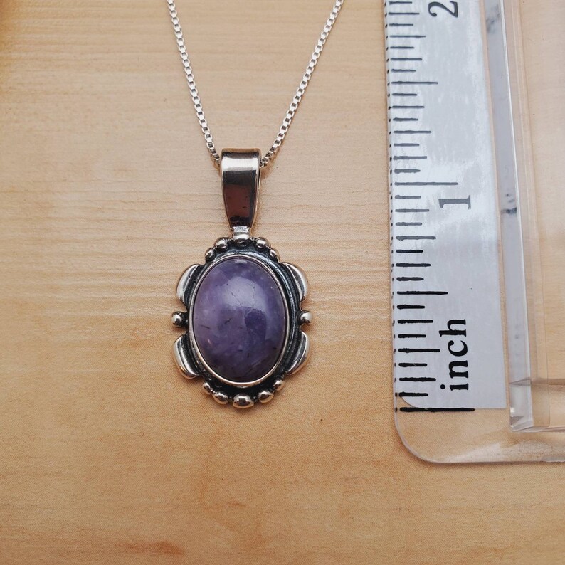 May include: A silver chain necklace with a purple oval gemstone pendant. The pendant is set in a silver frame with a decorative design. The necklace is measured against a ruler.