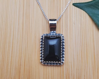 Può includere: Una collana con catena d'argento con un ciondolo in pietra nera. Il ciondolo è rettangolare e ha un bordo perlato.