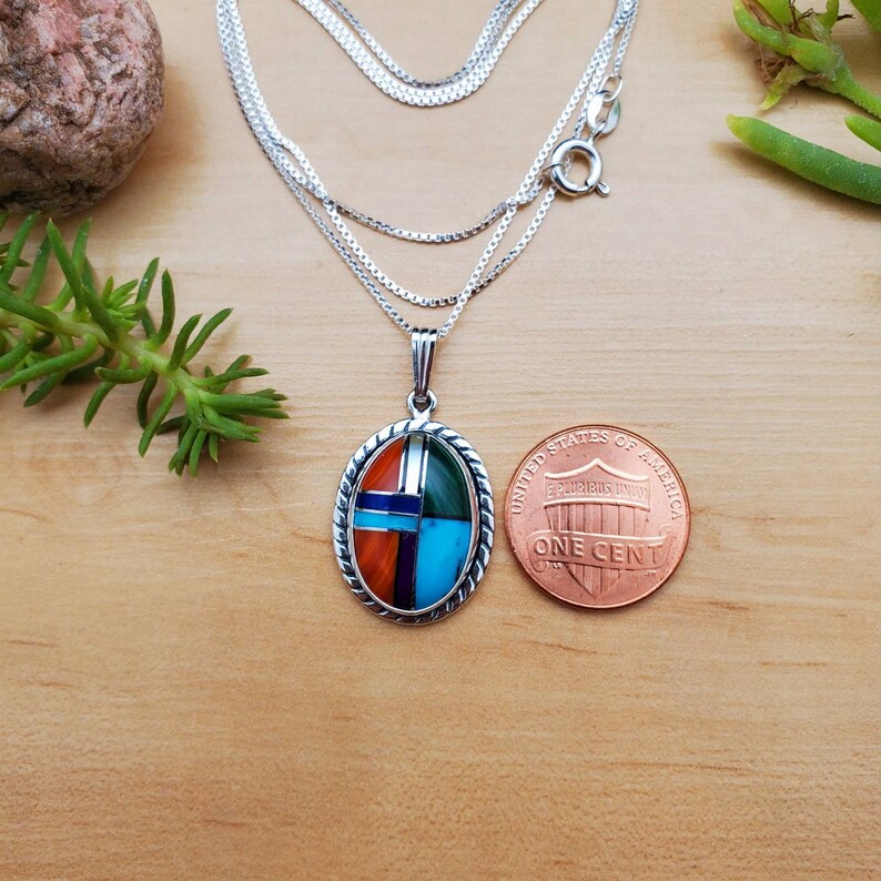 Pu&ograve; includere: Una collana con catena d'argento con un ciondolo ovale multicolore. Il ciondolo &egrave; realizzato con pietre intarsiate ed &egrave; grande circa come una moneta da un centesimo di dollaro.