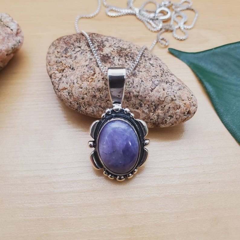 May include: A silver chain necklace with a pendant featuring a purple oval gemstone set in a silver bezel with a decorative border.