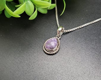May include: A silver necklace with a teardrop-shaped pendant. The pendant features a purple stone with a silver rope-like border. The necklace is on a black surface with green foliage in the background.