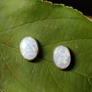 May include: A pair of silver stud earrings with oval-shaped white opal stones. The stones have a slight iridescent shimmer.