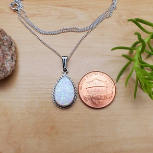 May include: A silver chain necklace with a teardrop-shaped pendant featuring a white opal stone. The pendant is surrounded by a silver border with a rope design. The necklace is shown next to a US penny for size comparison.