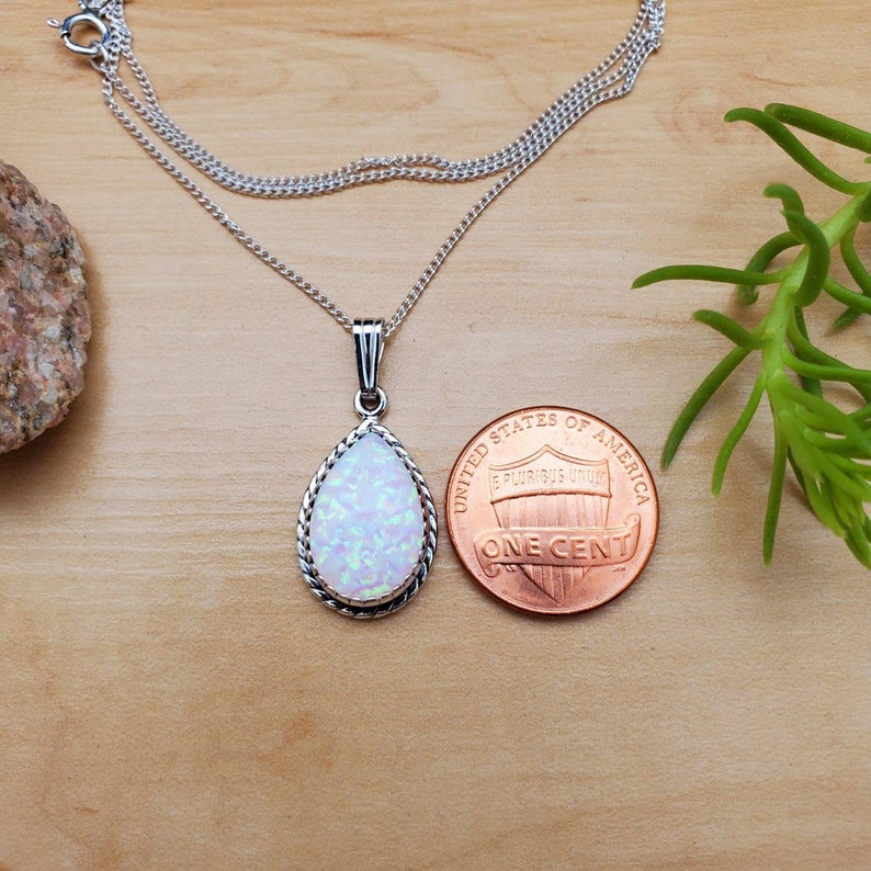 May include: A silver chain necklace with a teardrop-shaped pendant featuring a white opal stone. The pendant is surrounded by a silver border with a rope design. The necklace is shown next to a US penny for size comparison.