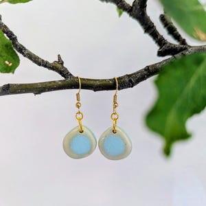 May include: A pair of drop earrings featuring small, rounded, pebble-shaped ceramic charms. Each charm has a light blue center surrounded by a smooth, off-white border. The charms are suspended from gold-toned metal fishhook ear wires.