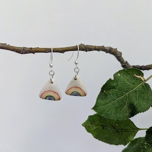 May include: A pair of dangle earrings featuring triangular off-white ceramic charms. Each charm displays a hand-painted rainbow motif with concentric arcs in blue, yellow, and red. The charms hang from silver-toned metal hooks and jump rings.