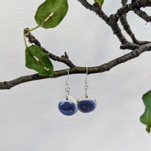 May include: A pair of drop earrings featuring smooth, rounded ceramic charms with a two-tone glaze. The top portion of each charm is white, transitioning into a deep, glossy blue at the bottom. The charms hang from silver-toned metal hooks.