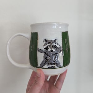 May include: White ceramic mug with a raccoon illustration. The raccoon is depicted in gray and black, with its arms outstretched. The mug has a green border on each side of the raccoon.