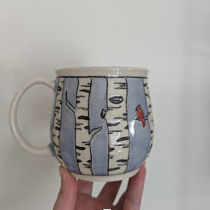 May include: A ceramic mug with a birch tree design. The mug features a light blue background with white birch tree trunks and black details. A red cardinal is perched on a branch. The mug has a white handle and a capacity of approximately 12 ounces.