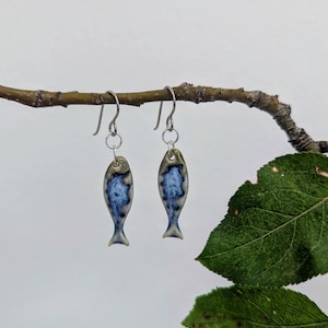 Puede incluir: Un par de pendientes en forma de pez con un acabado esmaltado suave en tonos crema, gris y azul oscuro. Cada pendiente cuelga de un sencillo gancho de metal plateado, mostrando un patrón moteado distintivo en toda la superficie.