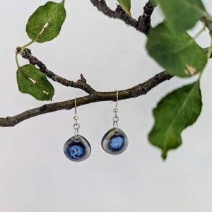 May include: A pair of drop earrings featuring small, rounded ceramic charms with a smooth, glossy finish. The charms have a dark blue center that fades into a light gray border. Each charm hangs from a silver-toned metal hook and jump ring.