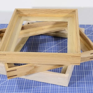 May include: Three wooden picture frames stacked on top of each other. The top frame is the largest and is made of a light brown wood. The middle frame is slightly smaller and is made of a darker brown wood. The bottom frame is the smallest and is made of a light wood.