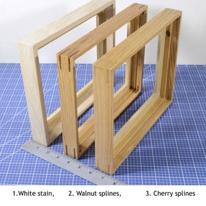 May include: Three wooden picture frames, each with a different finish. The first frame is stained white, the second frame has walnut splines, and the third frame has cherry splines.
