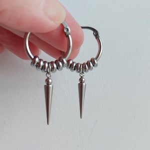 May include: A pair of silver hoop earrings with a series of small beads and a dangling, pointed cone-shaped charm. The earrings are made of a shiny, metallic material and are shown against a light background.