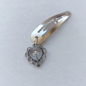 May include: Silver hair clip with a heart-shaped charm. The charm has a cross inside a heart-shaped frame, attached to a silver metal hair clip. The clip is designed to secure hair.