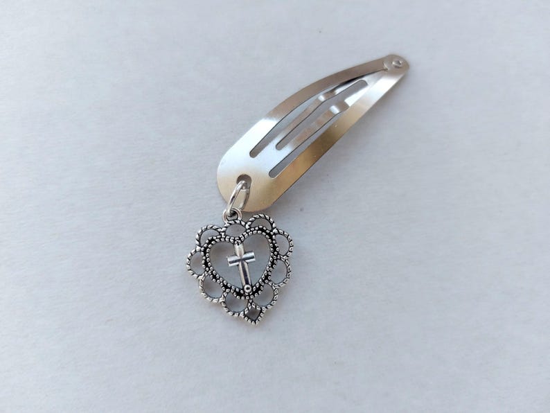 May include: Silver hair clip with a heart-shaped charm. The charm has a cross inside a heart-shaped frame, attached to a silver metal hair clip. The clip is designed to secure hair.
