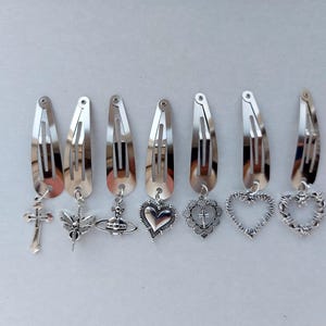 May include: A collection of silver hair clips, each adorned with a different charm. Charms include a cross, bee, heart, and planet. The clips are displayed in a line, highlighting their decorative nature.