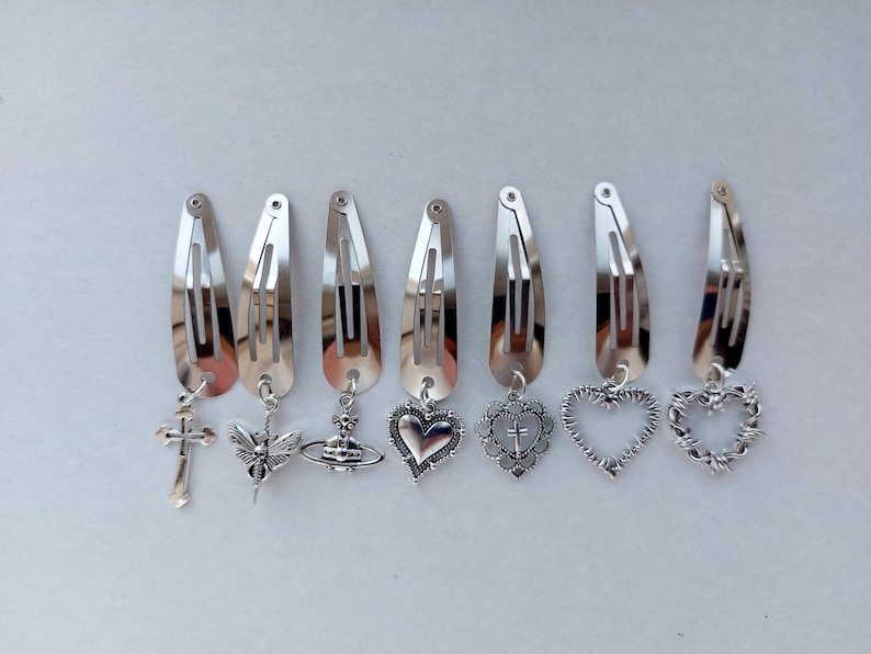 May include: A collection of silver hair clips, each adorned with a different charm. Charms include a cross, bee, heart, and planet. The clips are displayed in a line, highlighting their decorative nature.