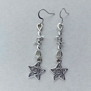 May include: A pair of silver-toned earrings. Each earring features a barbed wire design with a dangling star charm. The star charm has a spiral design in the center. The earrings hang from a simple hook.