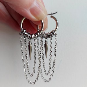May include: Silver hoop earrings with dangling chains and cone-shaped pendants. The earrings have a layered design with multiple chains and small beads around the hoops. The earrings are held up by a hand.