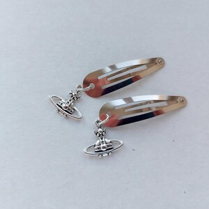 May include: Two silver hair clips, each with a planetary charm. The clips have a simple, streamlined design, with a small decorative charm hanging from each. The charms feature a detailed planetary design, adding a touch of elegance to the hair accessories.