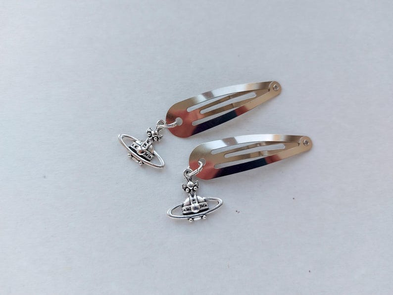 May include: Two silver hair clips, each with a planetary charm. The clips have a simple, streamlined design, with a small decorative charm hanging from each. The charms feature a detailed planetary design, adding a touch of elegance to the hair accessories.