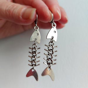 以下が含まれることがあります： 魚の骨の形をしたシルバー調のドロップピアス一対。各ピアスは、平らな頭部、突起のある肋骨を持つチェーンリンクの背骨、そして平らな尾部で構成されており、シンプルな金属製のフックで吊り下げられています。
