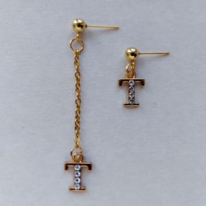 May include: A pair of gold-colored earrings. One earring is a stud with a gold letter "T" embellished with clear crystals. The other earring has a gold stud, a chain, and a dangling gold letter "T" with clear crystals.