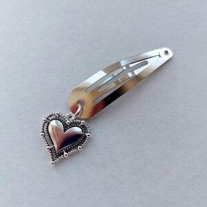 May include: Silver hair clip featuring a heart-shaped charm. The clip is a simple, rectangular design with a spring. The heart charm has a beaded edge and a smooth, polished centre. A charming accessory for any hairstyle.