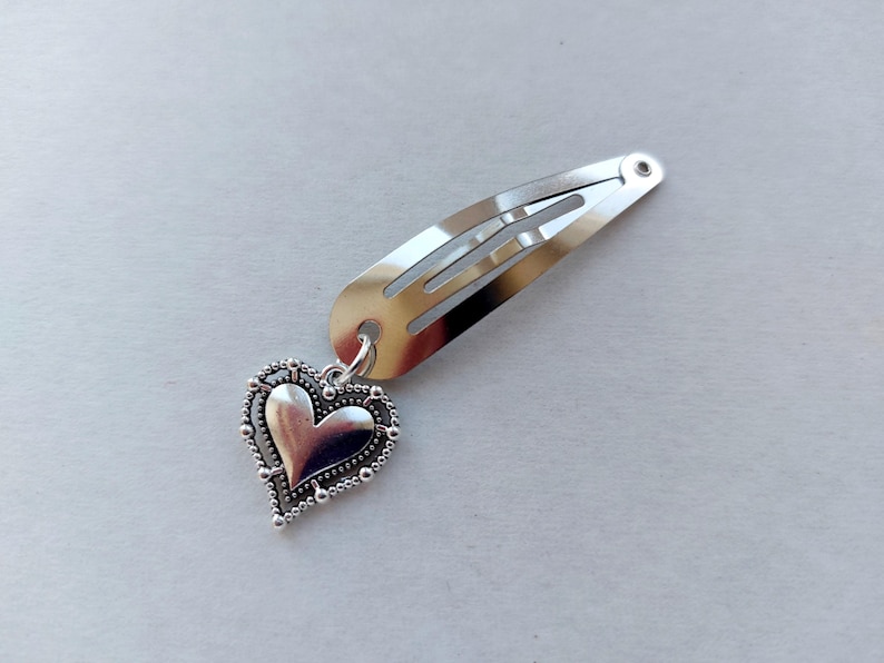 May include: Silver hair clip featuring a heart-shaped charm. The clip is a simple, rectangular design with a spring. The heart charm has a beaded edge and a smooth, polished centre. A charming accessory for any hairstyle.