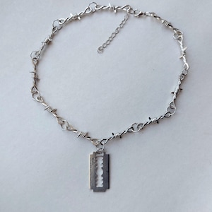 May include: A silver-toned necklace featuring a chain designed to resemble barbed wire, with a central pendant shaped like a rectangular razor blade. The necklace includes a small adjustable extender chain with a lobster claw clasp closure.