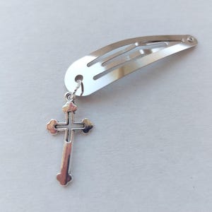 May include: A silver hair clip with a decorative cross charm. The cross pendant is detailed with rounded edges and a central cross design. The clip is a simple, curved barrette style, suitable for various hairstyles.