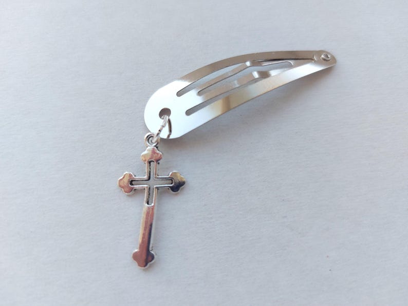 May include: A silver hair clip with a decorative cross charm. The cross pendant is detailed with rounded edges and a central cross design. The clip is a simple, curved barrette style, suitable for various hairstyles.