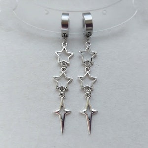 Pode incluir: Um par de brincos de prata com três pingentes em forma de estrela pendurados em cada brinco. Os brincos são feitos de aço inoxidável e têm um design de aro.