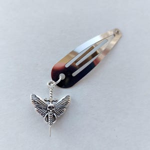 May include: Silver-coloured hair clip with a decorative pendant. The pendant features a detailed moth design with a skull face, wings, and a sword. The hair clip is approximately 5 cm long.