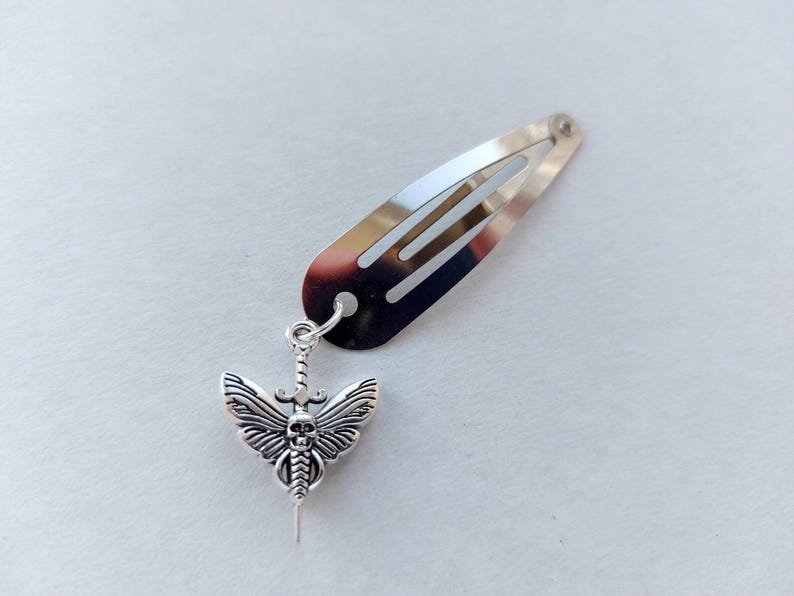 May include: Silver-coloured hair clip with a decorative pendant. The pendant features a detailed moth design with a skull face, wings, and a sword. The hair clip is approximately 5 cm long.