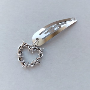 May include: Silver hair clip featuring a heart-shaped charm. The charm is made from silver-coloured wire, formed into a heart, and connected to the clip. The clip is a standard barrette style, made of shiny metal.