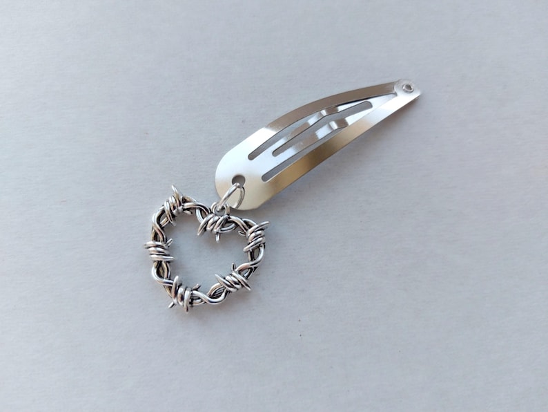 May include: Silver hair clip featuring a heart-shaped charm. The charm is made from silver-coloured wire, formed into a heart, and connected to the clip. The clip is a standard barrette style, made of shiny metal.