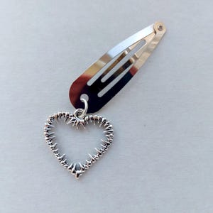May include: Silver hair clip with a heart-shaped charm. The clip is a barrette style, and the heart charm has a textured, spiky design. The clip and charm are made of a shiny, metallic material.