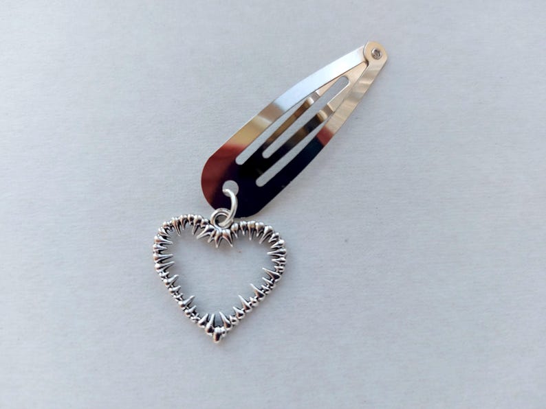 May include: Silver hair clip with a heart-shaped charm. The clip is a barrette style, and the heart charm has a textured, spiky design. The clip and charm are made of a shiny, metallic material.